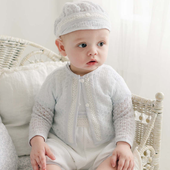 A baby, dressed in the Harrison Blue Knit Sweater and a matching hat, sits on a white wicker chair with cushions. The baby gazes slightly to the side in a well-lit room adorned with sheer white curtains, creating an overall soft and serene ambiance.