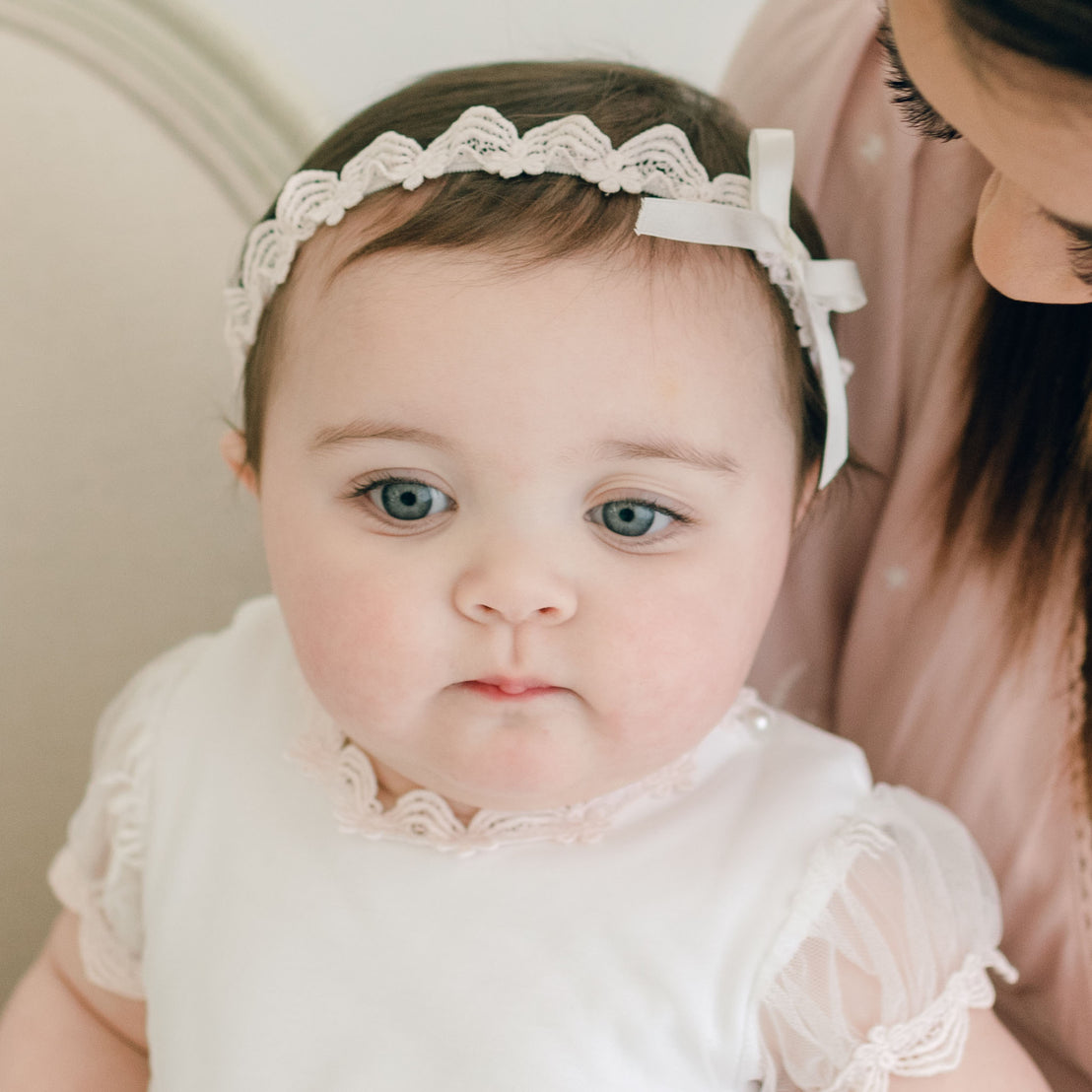 A baby wearing the Joli Lace Headband looks up, while a woman, partially shown, leans in close to the baby.