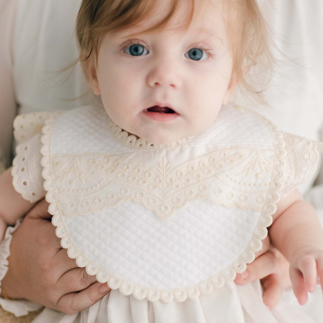 Baby wearing the Ingrid Bib crafted with a soft quilted cotton, accented with a sweet scalloped ecru edge lace and detailed with the matching Ingrid eyelet lace