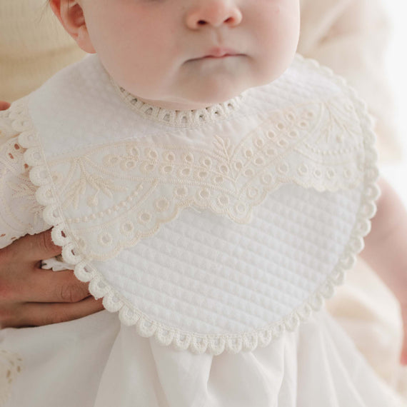 A baby wearing the Ingrid Bib, featuring intricate lace detailing around the edges and paired with a matching outfit. The quilted cotton of the bib adds a vintage touch. With a calm expression, the baby is gently supported by an adult's hand from behind.