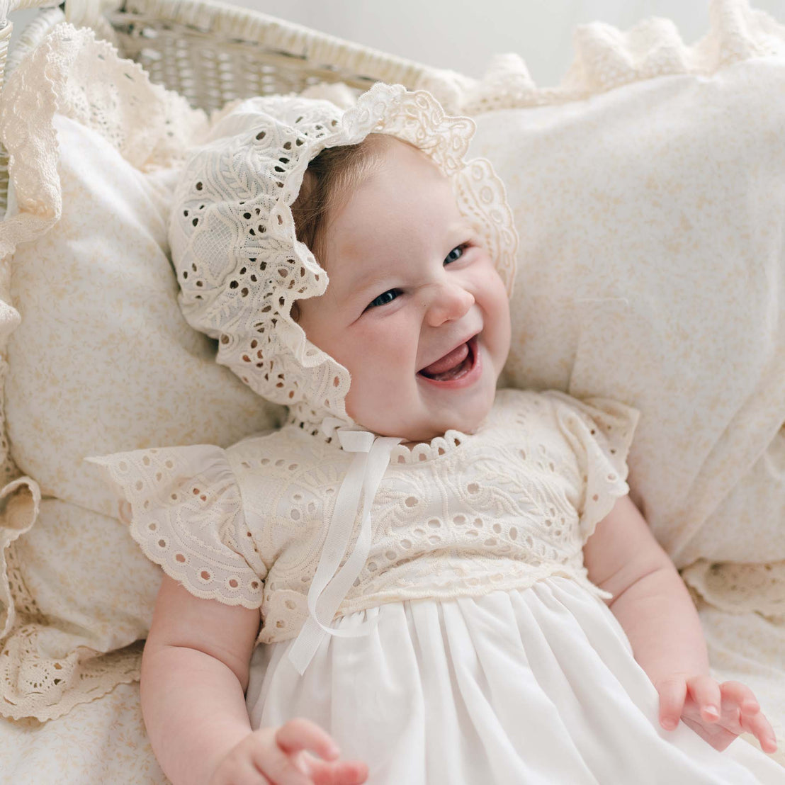 A smiling baby dressed in a white, lacy Ingrid Tiered Gown & Bonnet with eyelet details is lying down on a matching lacy pillow. The baby has a joyful expression and is surrounded by light-colored, delicate fabrics, perfect for her Girls Baby Blessing.