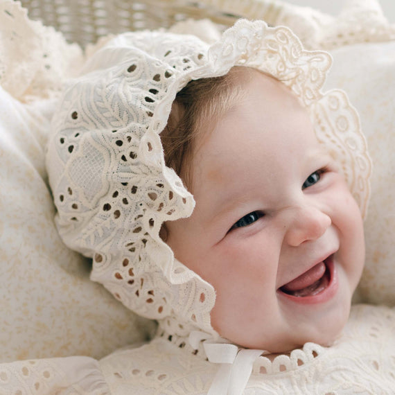 A baby wearing a delicate Ingrid Bonnet and matching Christening Gown is smiling joyfully. The intricate lace patterns add elegance, and the baby appears to be resting on a soft, light-colored cushion. Handmade in the USA, the scene exudes warmth and happiness.