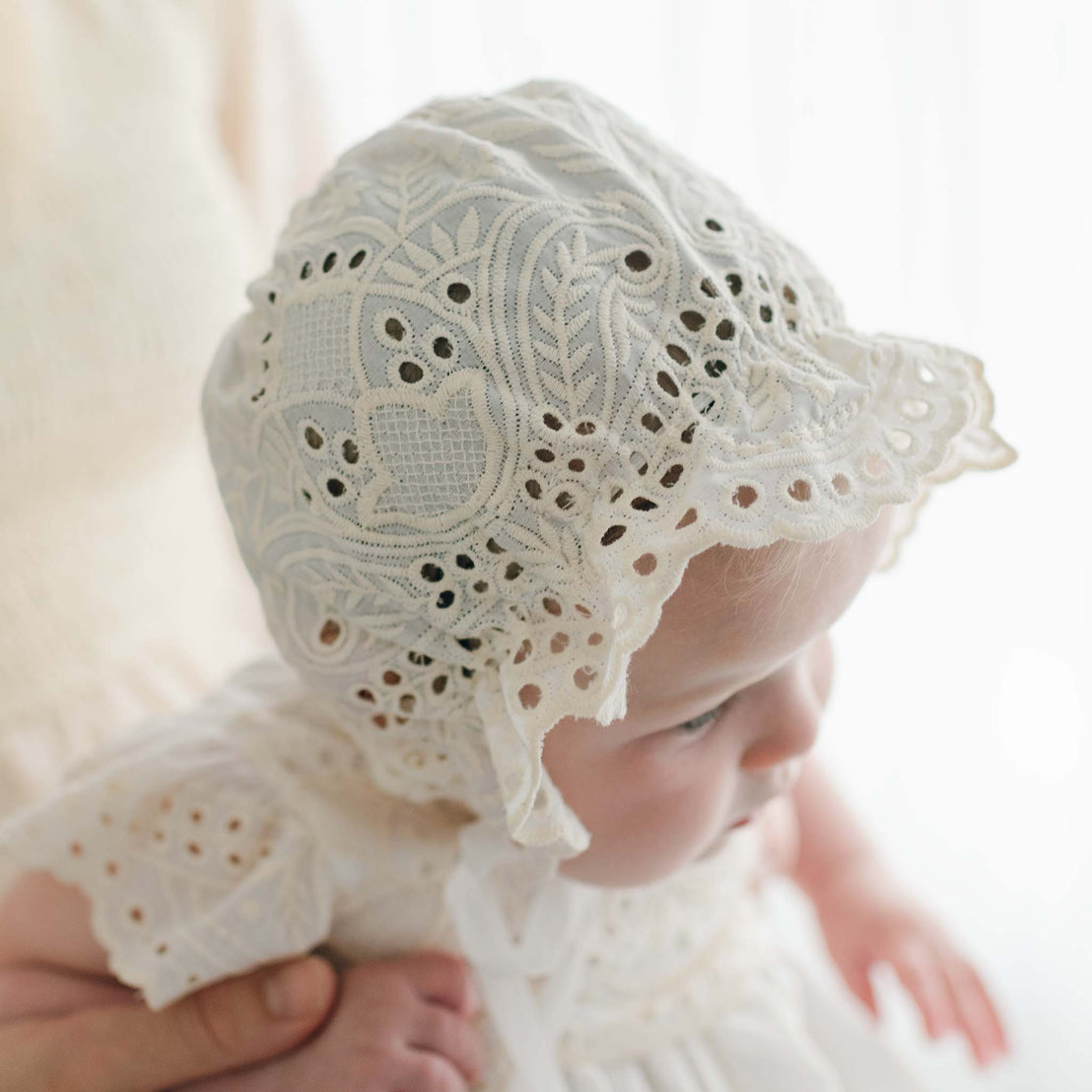 A baby wearing an Ingrid Tiered Gown & Bonnet, adorned with intricate lace details, is being held by a partially visible adult. The softly blurred and bright background emphasizes the baby and the exquisite craftsmanship of the lacework.