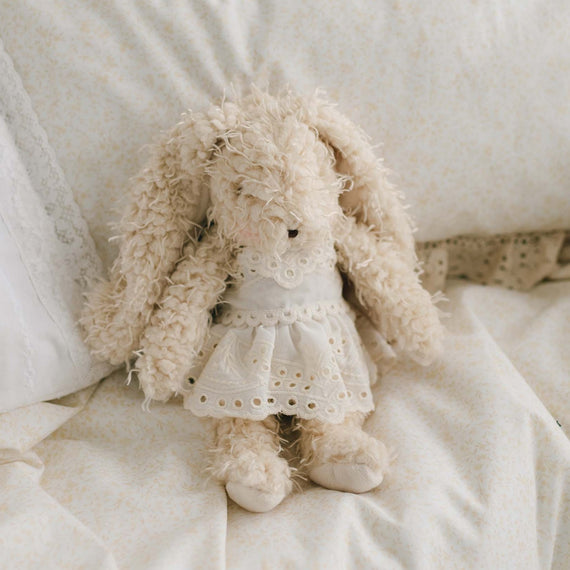 Ingrid Bunny, a fluffy, cream-colored soft bunny with an embroidered face, sits on a soft white bed adorned with floral-patterned linens. Dressed in a delicate white lace dress, Ingrid Bunny enhances the cozy setting highlighted by lace accents on the pillows and bedding, making it a perfect gift for a baby.