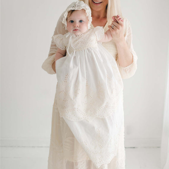 A woman holding a baby dressed in the Ingrid Tiered Gown & Bonnet, which features natural embroidery and intricate detailing. The woman is wearing a sheer, light-colored dress; her face is only partially visible. The background is plain white.