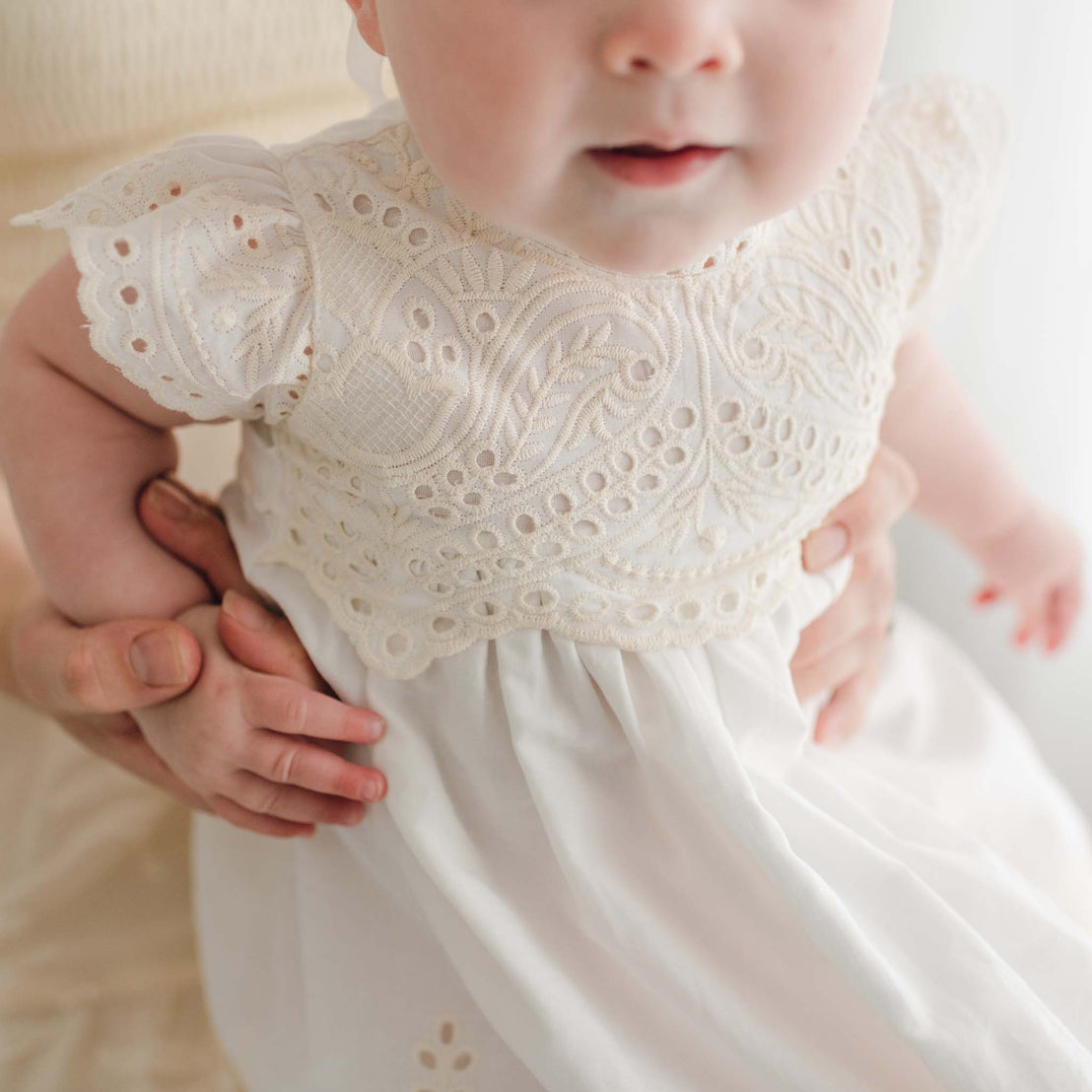 A baby wearing a delicate white lace Ingrid Tiered Gown & Bonnet is gently supported by an adult's hands. The gown features intricate lace detailing on the bodice and sleeves. The baby is viewed from the chest up, with part of the adult's hand visible.