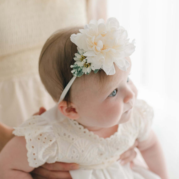 A baby wearing a white lace dress and the Ingrid Flower Headband in ivory dahlia looks to the side while being held. The soft lighting and light-colored background add to the delicate, serene atmosphere of the image, enhancing a sense of baby comfort.