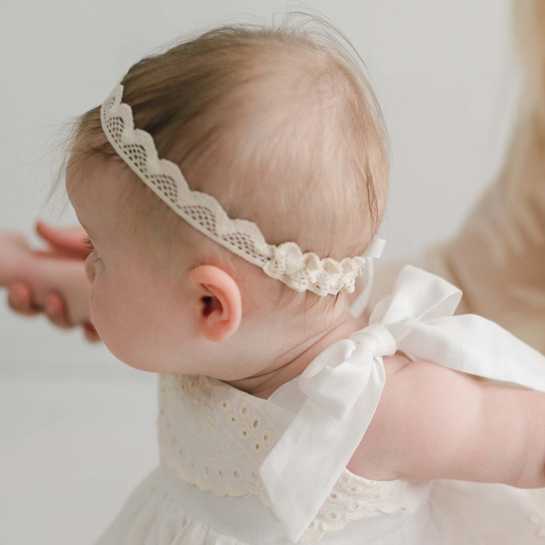 A baby with light skin and fine hair, wearing the delicate handmade Ingrid Lace Baby Headband and a white christening gown adorned with a large bow on the shoulder, is seen from the side. An adult's hand gently holds the baby's arm, providing support. The background is minimalistic and softly focused.