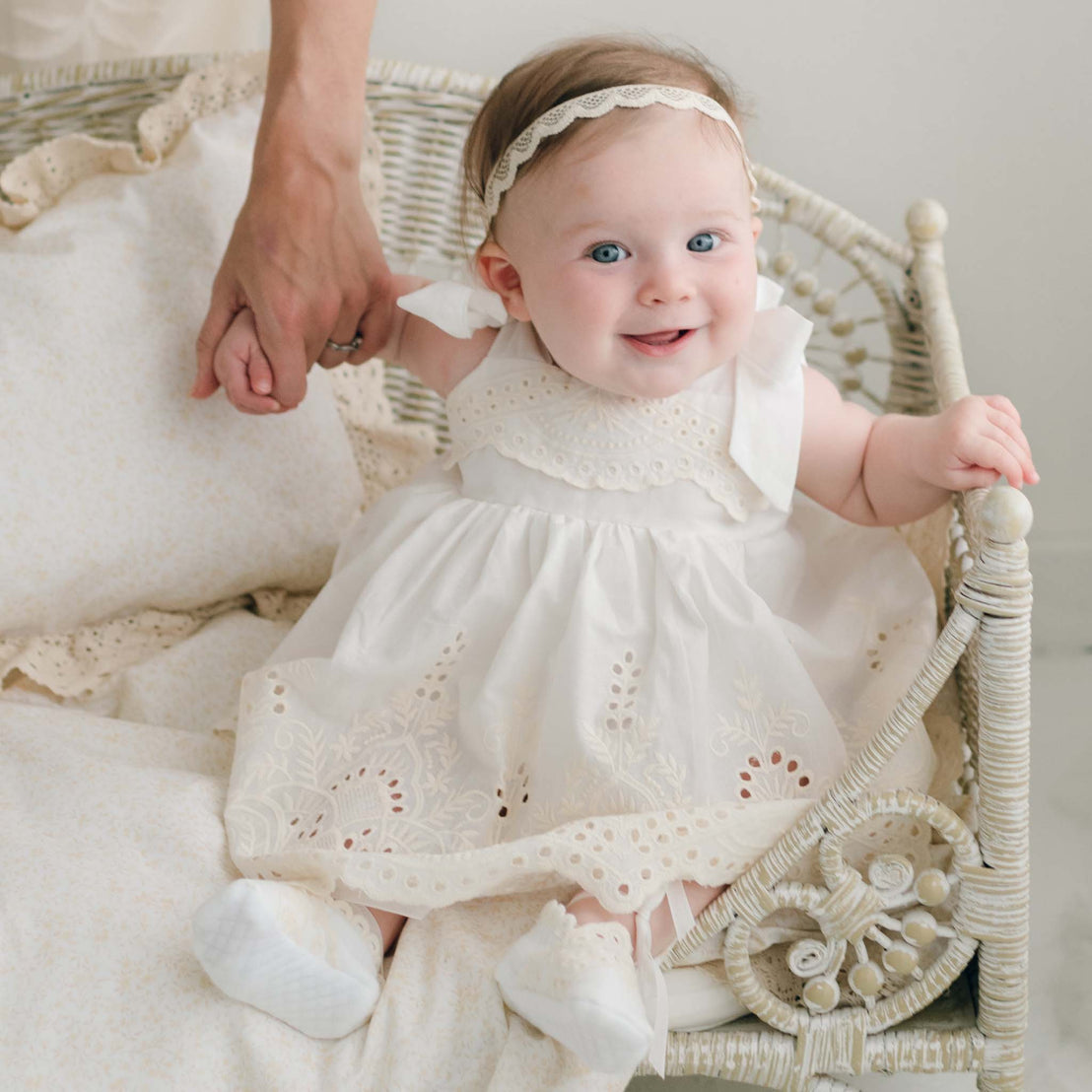 A smiling baby dressed in the Ingrid Romper Dress and a headband sits comfortably in a decorative wicker chair. An adult's hand gently holds onto the baby's for support, with neutral, cozy bedding in the background accentuating the intricate details of her handmade outfit.