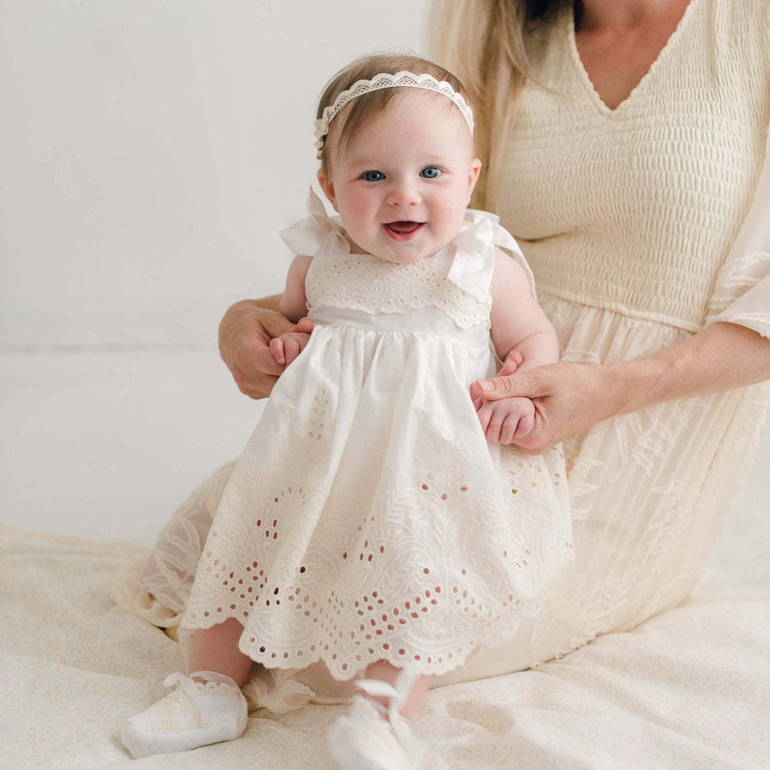 A cheerful baby girl, dressed in the Ingrid Romper Dress complemented by matching shoes and a headband, is sitting on a woman's lap who dons a light-colored dress. The background is bright and neutral.