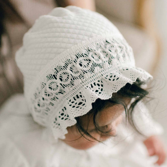 A baby girl with dark hair wearing the Lily Quilted Cotton Bonnet made with ivory quilted cotton, light ivory stretch lace, and light ivory cotton ribbon. The baby is looking down away from the camera.