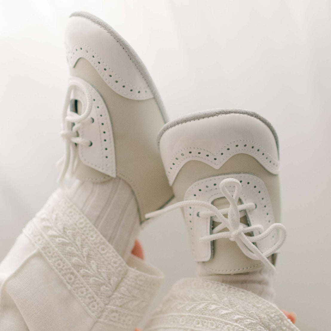 A close-up of baby feet wearing white, handmade Ivory Two Tone Wingtip Shoes with decorative perforations. The baby is dressed in a cream outfit with intricate lace patterns, positioned against a soft, light background.