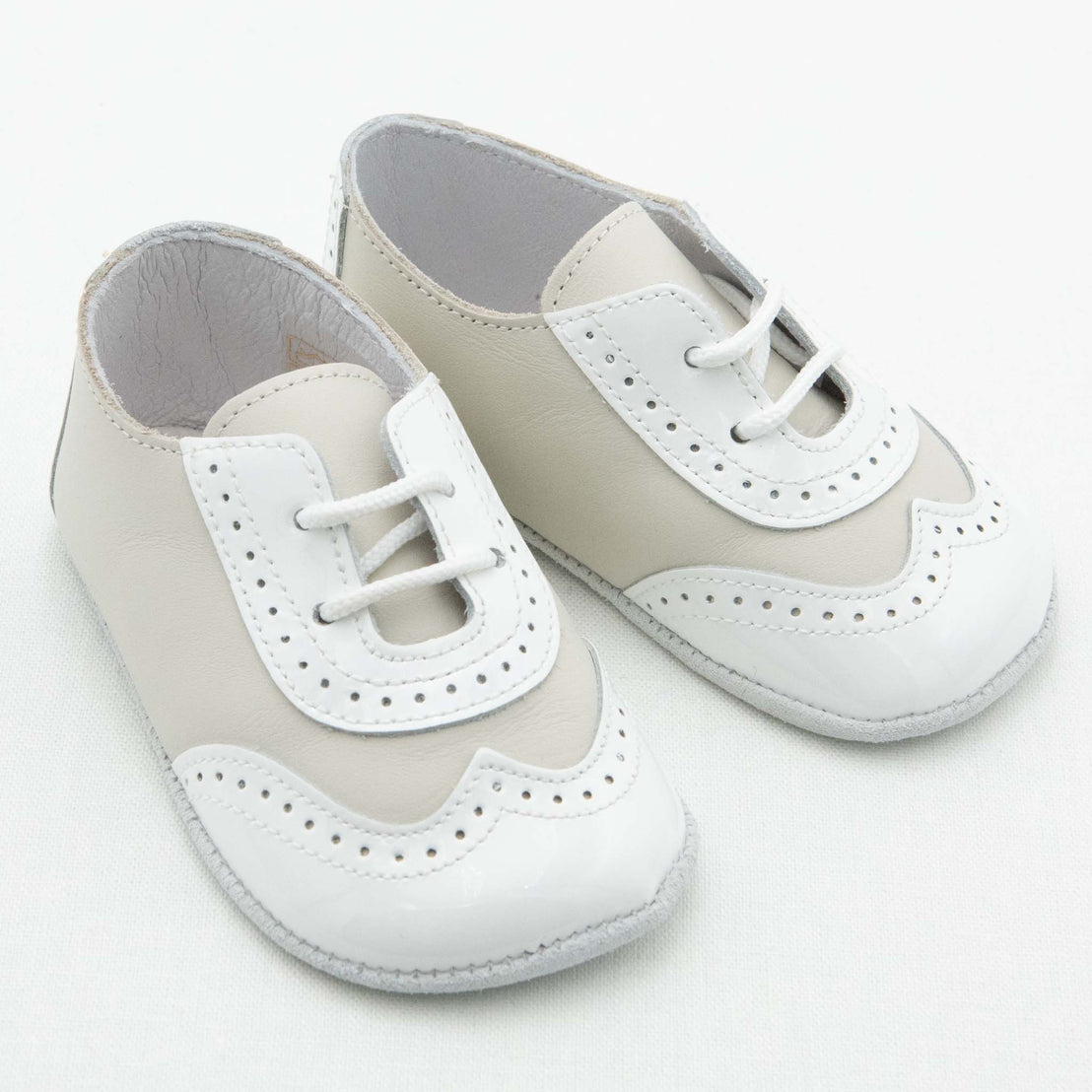 The Ivory Two Tone Wingtip Shoes are a pair of small, elegant footwear in a combination of white and light beige. These handmade leather shoes feature lace-up fronts with decorative perforations and a wingtip design, giving them a classic, refined look. They have soft soles suitable for infants.