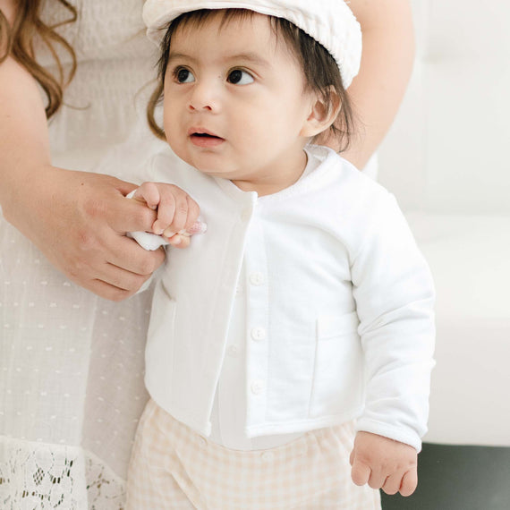 A baby with expressive eyes, dressed in an Ian French Terry Jacket and traditional patterned pants, holds an adult's hand, looking up with curiosity. The adult is partially visible.