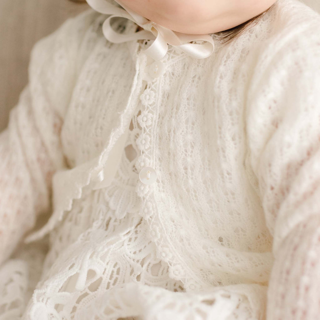 Close-up of the Lola Knit Sweater, featuring soft light ivory knit fabric, button closures, and delicate lace trim, layered over a lace christening gown.