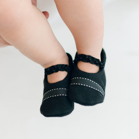 Close-up of a baby's legs and feet wearing James Booties, black boys booties with white stitching details. Handmade in the USA, these booties feature small ruffles around the ankle. The background is softly blurred, highlighting the baby's legs and footwear.