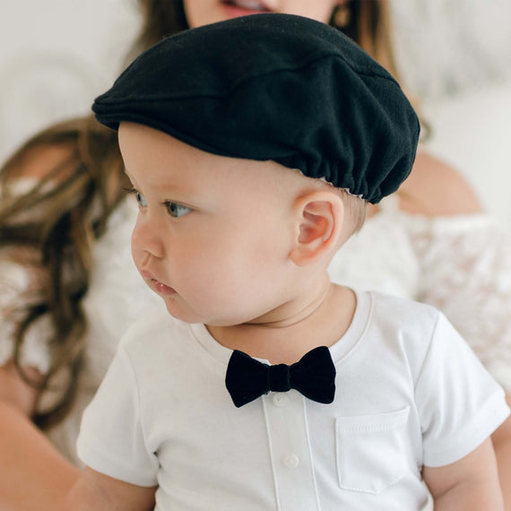 A baby wearing a white quilted cotton outfit and a black bow tie is looking to the side while also sporting the James Newsboy Cap in black. In the background, an adult is partially visible but out of focus.