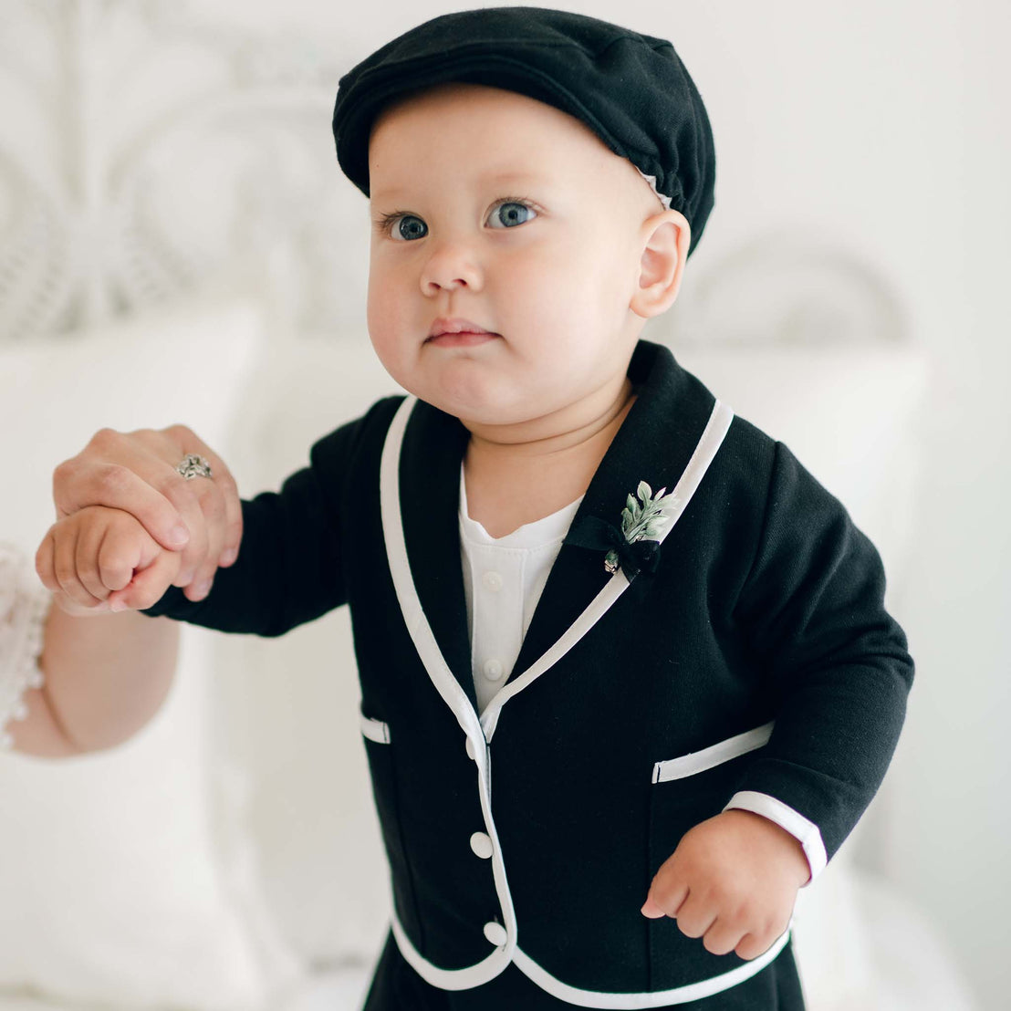 A baby in the James 3-Piece Suit, featuring formal black attire with Dupioni silk trim and a matching black cap, holds an adult's hand for support. The baby gazes ahead with a serious expression. The background is softly blurred and white, with subtle decorative elements that enhance the elegance of this handmade baby boy suit.