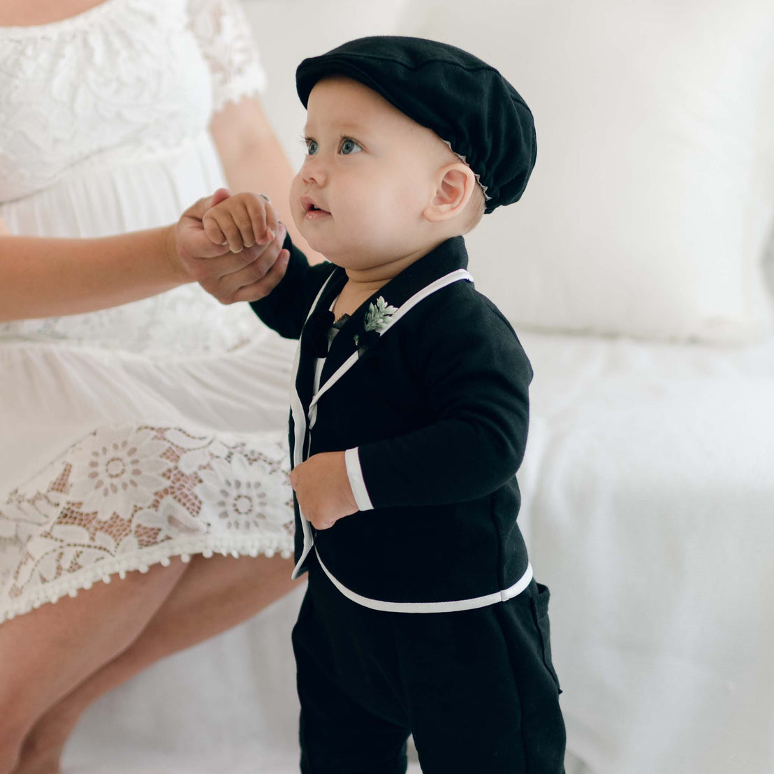 A toddler wearing the James 3-Piece Suit, featuring delicately crafted Dupioni silk trim and a matching black flat cap, holds an adult's hand. The child stands on a white surface, with part of an adult in a white lace dress visible in the background. The scene appears to be indoors.