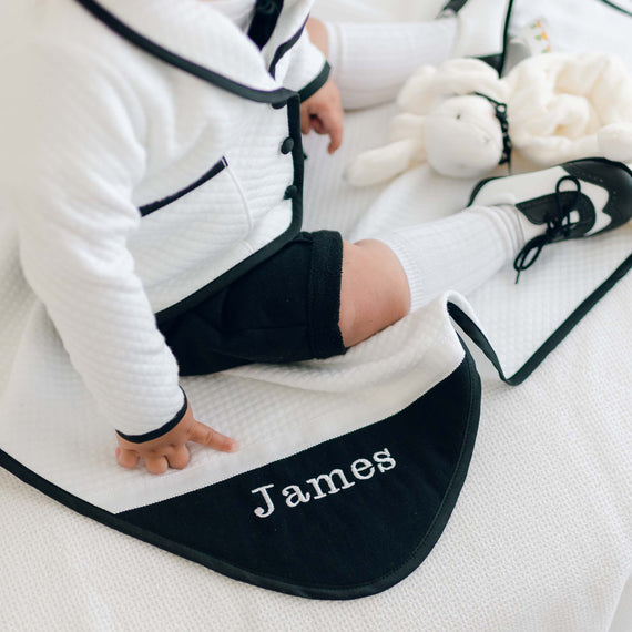 A young child dressed in a formal outfit sits on the James Personalized Blanket, which features an elegant white and black design. The child's finger points to the corner of the handmade blanket where "James" is beautifully embroidered. A white stuffed animal can be seen in the background, making this a perfect personalized gift for any occasion.