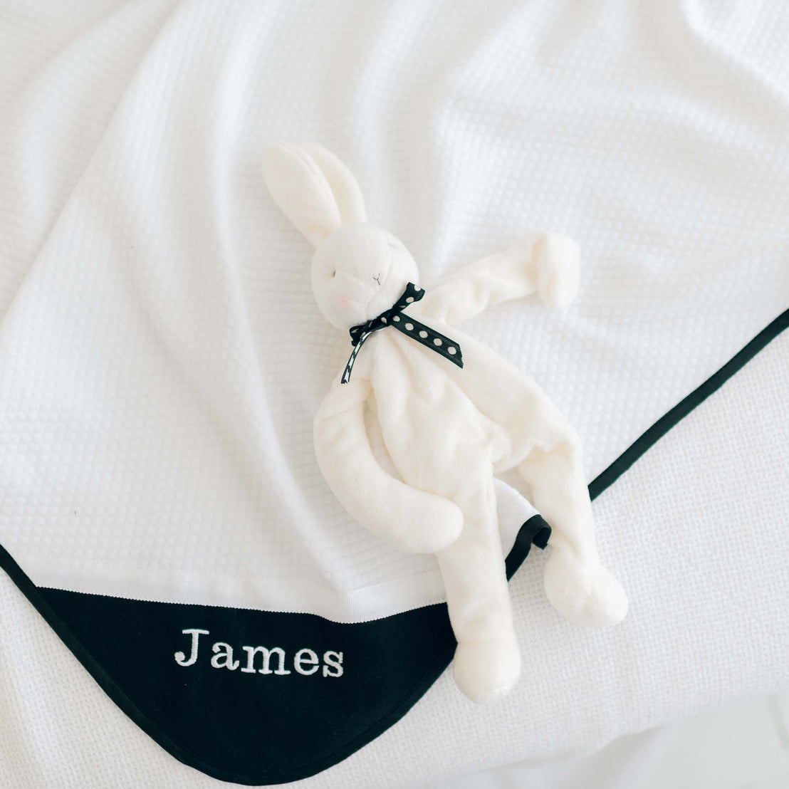 A soft, white stuffed bunny with a black polka dot ribbon around its neck is lying on a James Personalized Blanket. The blanket features a black border and is embroidered with the name "James" in white letters, making it a perfect personalized baby shower gift.