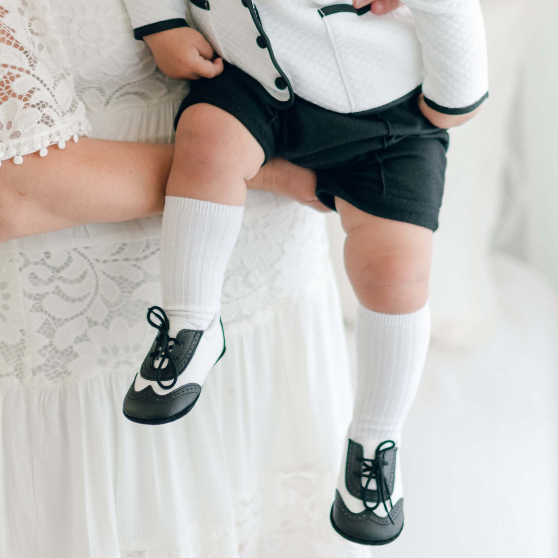 The lower half of a baby boy being held by his mother. He is wearing the James-3 Piece Suit and matching Black and White Leather Wingtip Shoes (with ribbed knee socks).