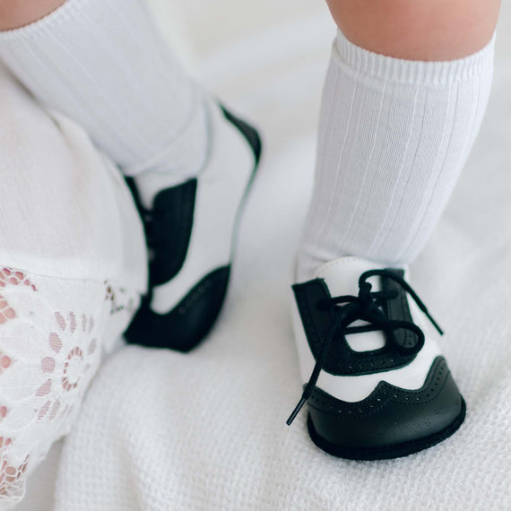 The legs of a baby boy standing on white textured fabric. He is wearing Black and White Leather Wingtip Shoes with matching White Ribbed Knee Socks.