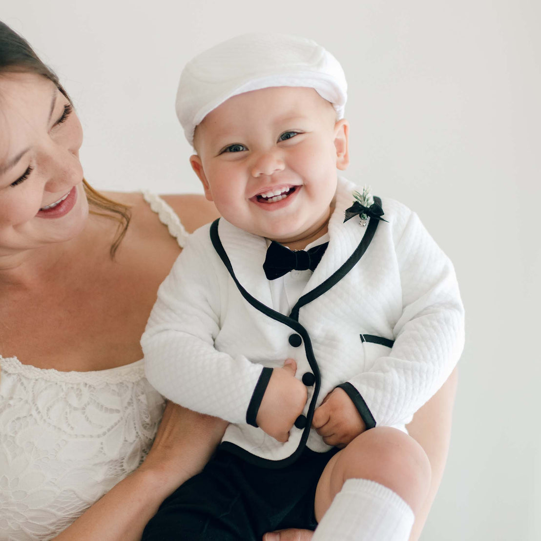 A smiling baby dressed in the James 3-Piece Suit, complete with Dupioni silk trim, a black bow tie, and a white cap, is being held by a woman in a white dress. The baby is looking directly at the camera with a happy expression.