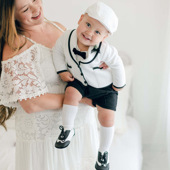 A woman with long hair in a white lace dress holds a smiling baby boy who is dressed in the James 3-Piece Suit. This outfit includes a handmade suit with Dupioni silk trim, featuring a sophisticated white hat, a stylish white jacket with black accents, an elegant bow tie, black shorts, white knee-high socks, and black and white shoes.