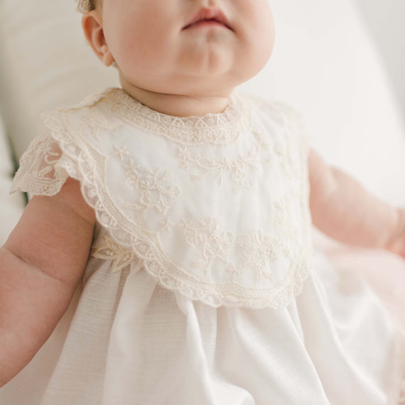 A close-up image of a baby wearing a delicate white Jessica Cotton Bib with champagne lace detailing around the collar and sleeves. Made from 100% ivory cotton, the baby is seated, with a focus on the upper part of the bib and chubby cheeks visible. The background is softly blurred.