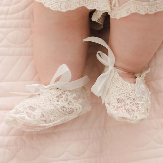 Close-up of a baby's feet wearing delicate, off-white Jessica Lace Booties adorned with ribbons on a soft, quilted, pink blanket. The baby's legs are partially visible, with a hint of champagne lace and floral appliqué on their lacy outfit above the booties.