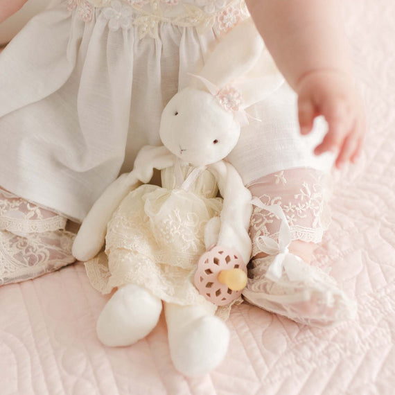 A baby in a white lace dress sits on a pink quilted blanket, reaching out with one arm. Next to her is a Jessica Silly Bunny Buddy | Pacifier Holder in a matching lace dress, clutching a small donut-shaped toy. Both the baby and the bunny's dresses feature delicate floral lace details.