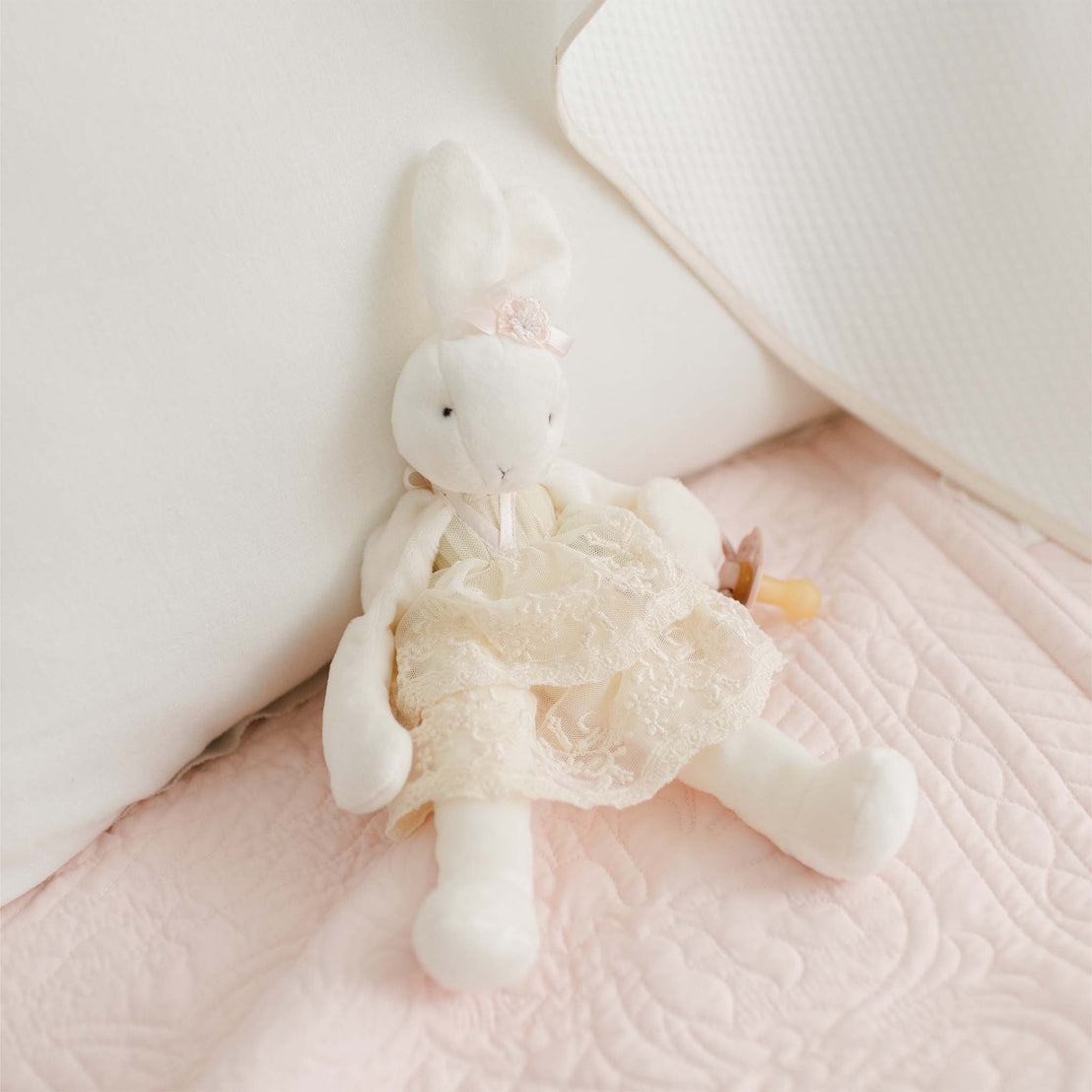 A Jessica Silly Bunny Buddy | Pacifier Holder, a plush white bunny doll wearing a lace dress and a pink flower headband, which doubles as a soft velcro pacifier holder, sits against a white pillow on a soft, light pink quilted blanket.