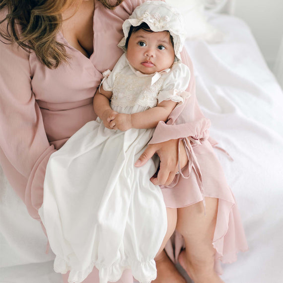 A baby girl dressed in a Jessica Newborn Gown & Bonnet Set is held on the lap of a woman in a pastel pink floral applique dress. The baby looks up with a calm expression, and the woman’s partial face is visible, both seated on a white fabric background.