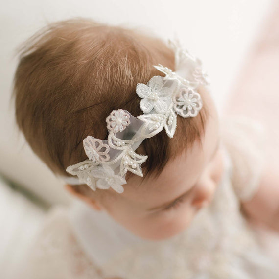 A baby with brown hair is wearing a delicate, white Jessica Beaded Flower Headband. The baby is dressed in a white, lacy outfit and is gazing downwards. The background is softly blurred, focusing attention on the baby's head and details of the accessories.