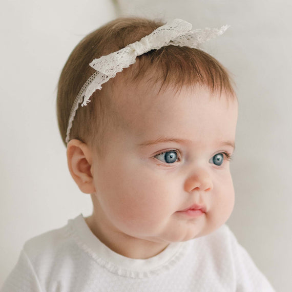 A baby with blue eyes is dressed in a white outfit and wears a Hailey Lace Headband with a delicate lace bow. The baby looks off to the side against a plain light background.