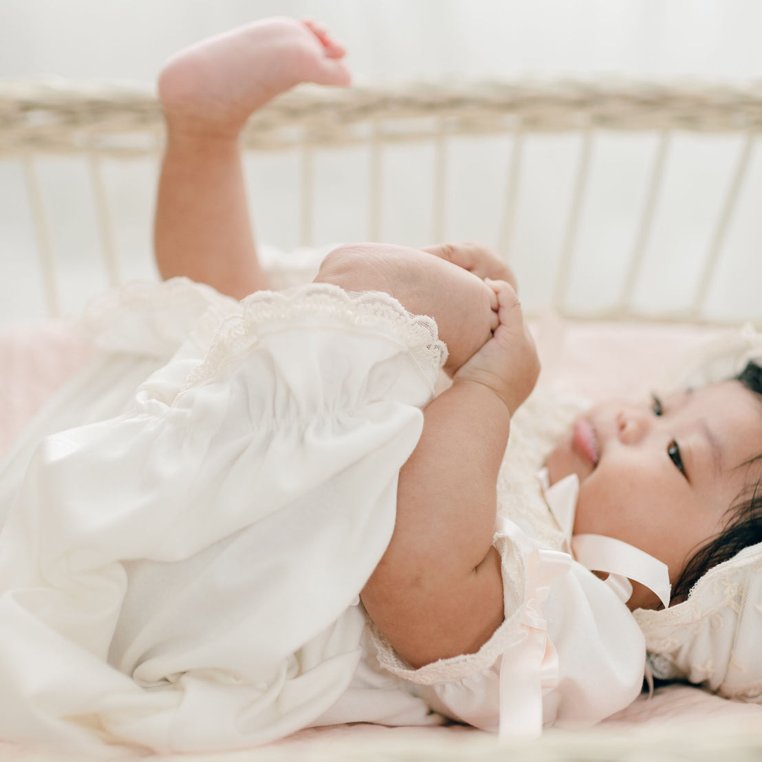 A baby dressed in a white Jessica Newborn Gown & Bonnet Set lies in a wicker crib, playing with their foot. The baby's chubby cheeks and relaxed expression suggest a calm and playful moment. The background is light and softly focused.