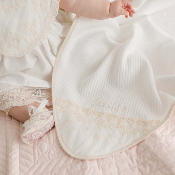 A baby's legs and feet are shown wearing delicate lace booties and a white outfit. The baby is draped in a Jessica Personalized Blanket with intricate lace detailing and the name "Jessica" embroidered on it. The serene scene is set on a pale pink cotton quilted blanket, adding to its soft charm.