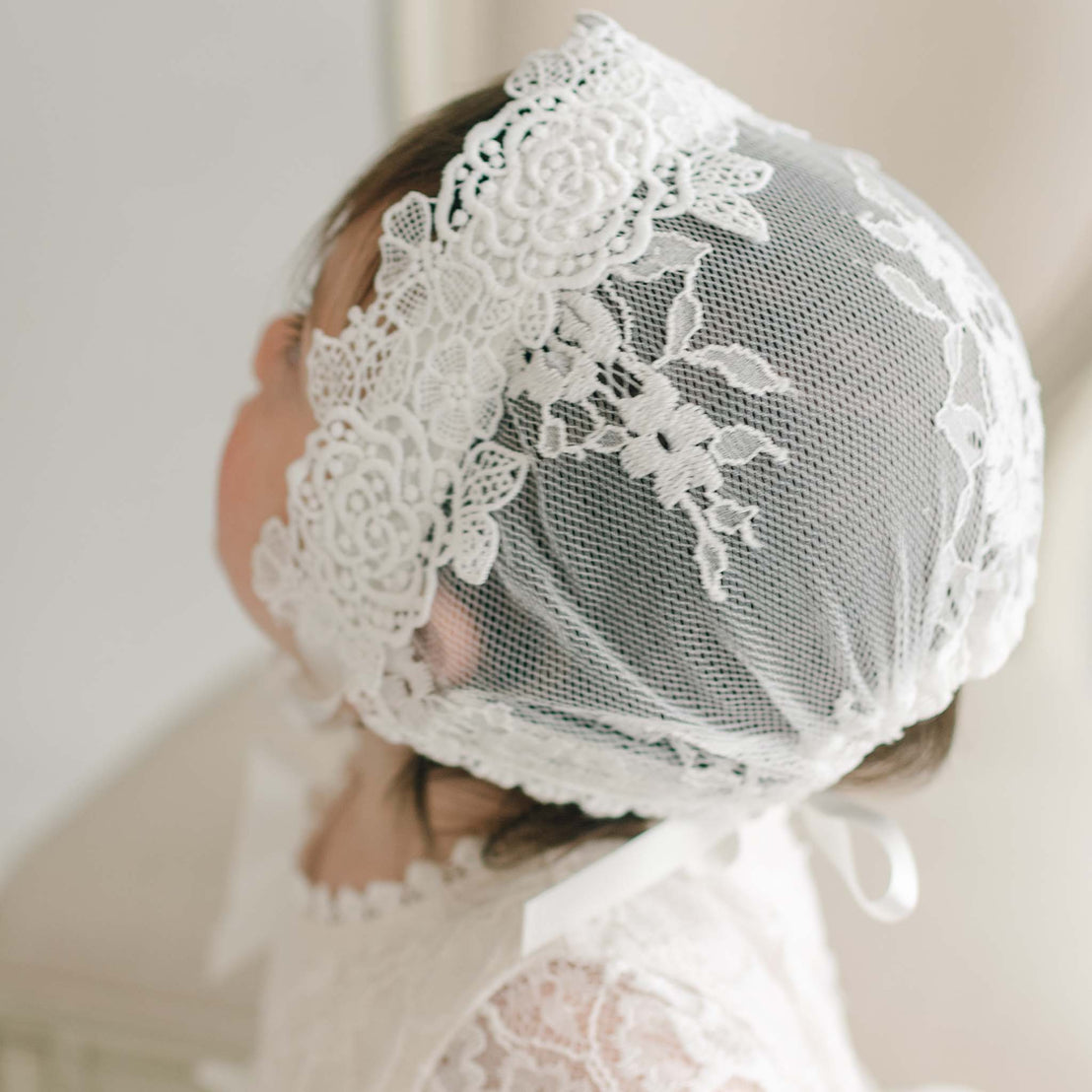 A close-up image of a baby wearing a delicate Juliette Lace Bonnet. The bonnet, handmade in the USA, features intricate floral patterns and covers the baby's head, while ivory silk ribbons tie it under the chin. The baby is turned slightly to the side, showcasing the detailed design of the bonnet.