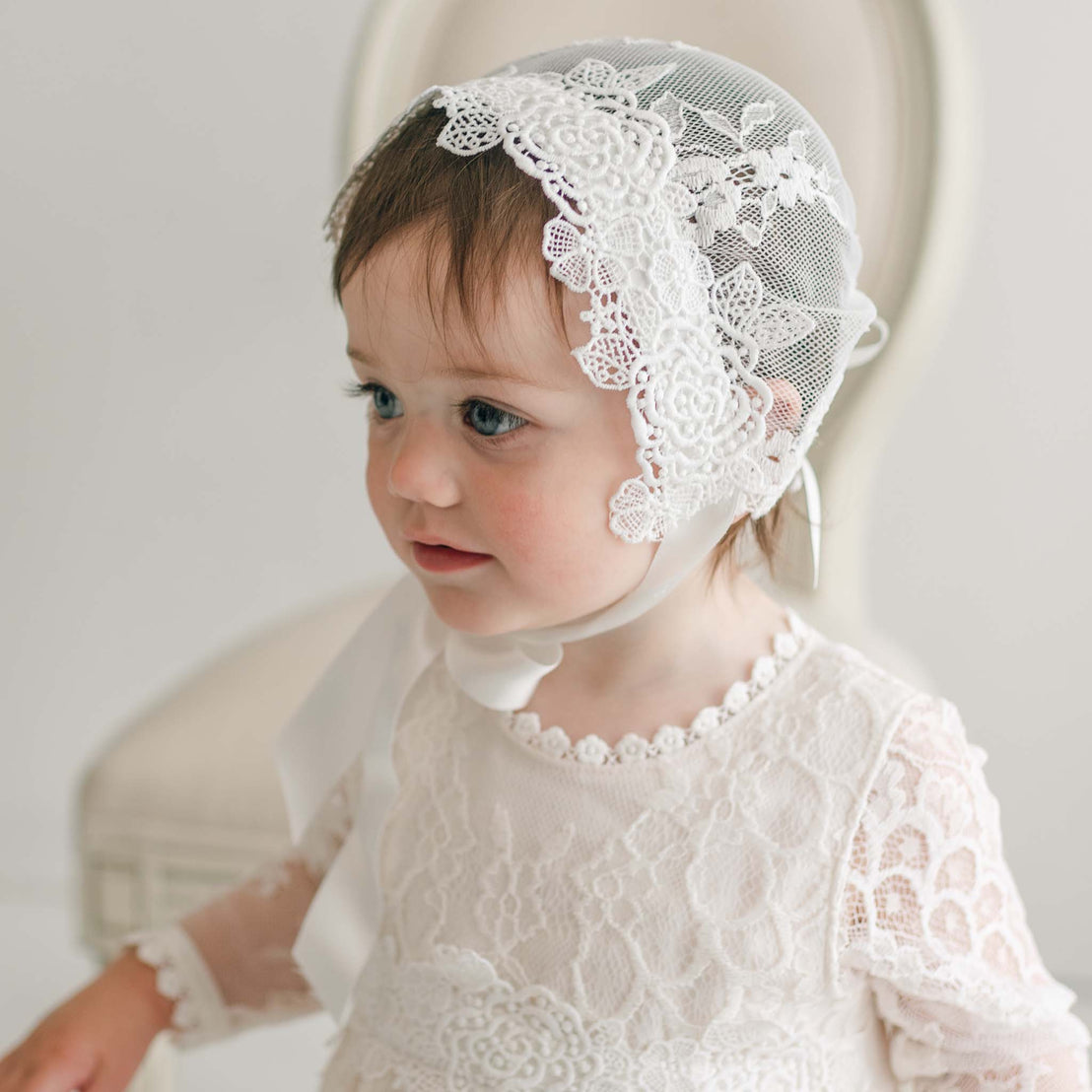 A young child with blue eyes wears a Juliette Lace Bonnet adorned with intricate lace patterns and ivory silk ribbons, paired with a white lace dress featuring delicate floral details. Sitting in a softly lit room, the child is framed by the grace of a cream-colored chair, embodying timeless elegance.