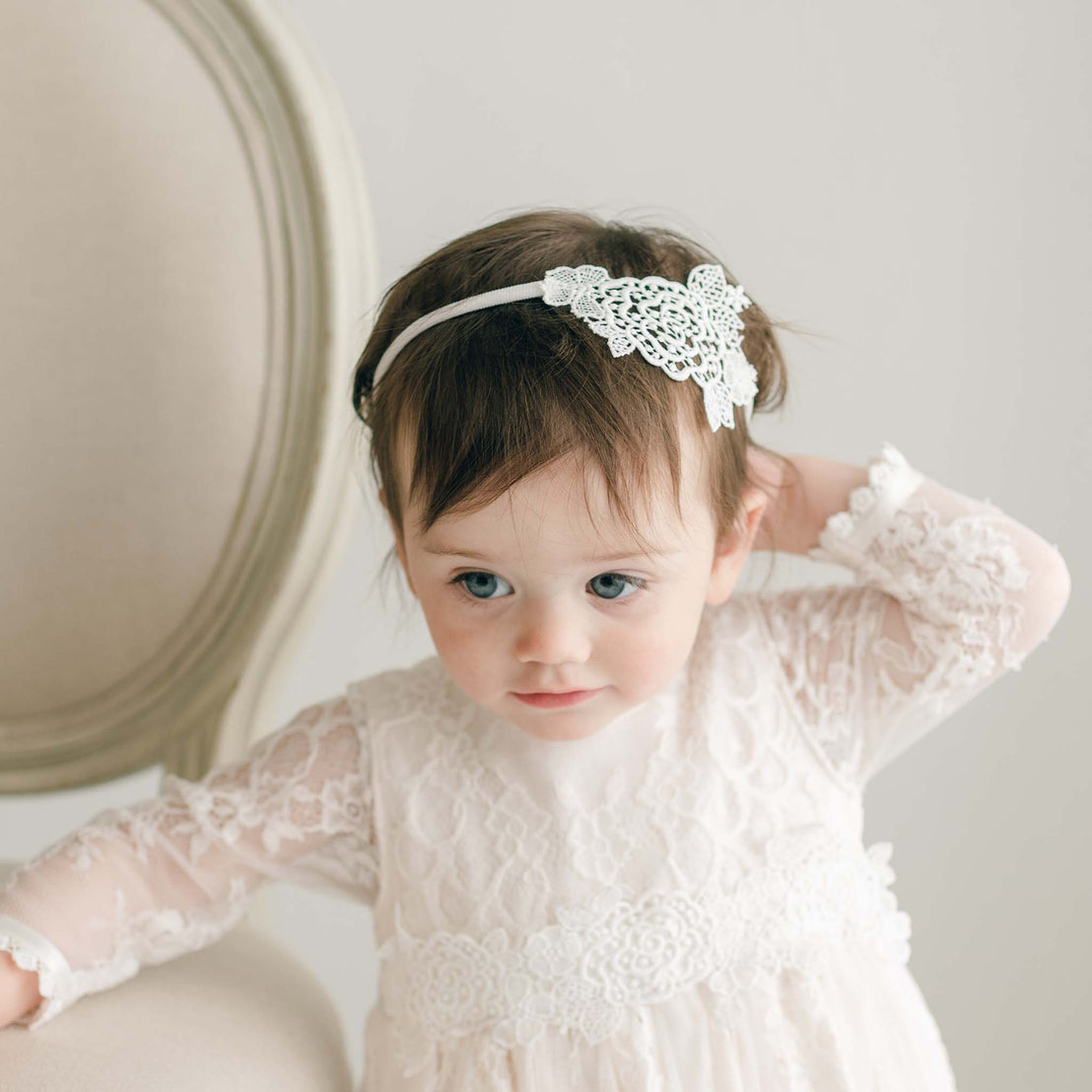 A young child with brown hair, adorned in a white lace dress and the elegant Juliette Lace Headband in light ivory rose, is seated on a beige chair. With one hand behind their head, the child gazes thoughtfully to the side against a plain, light-colored background.