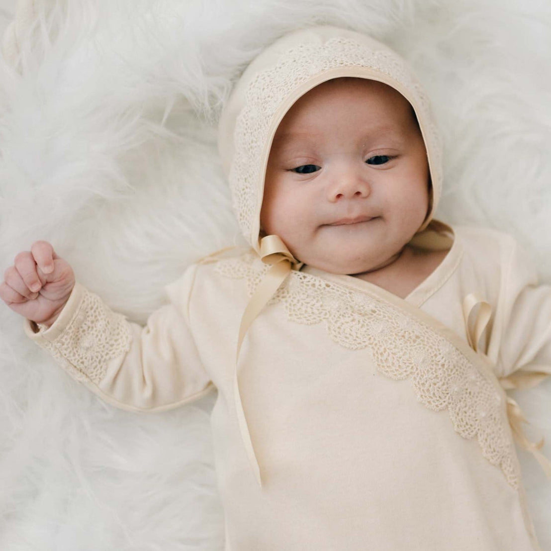 A serene newborn girl wearing the Mia Knot Gown & Bonnet lies on a white furry blanket, giving a gentle smile.