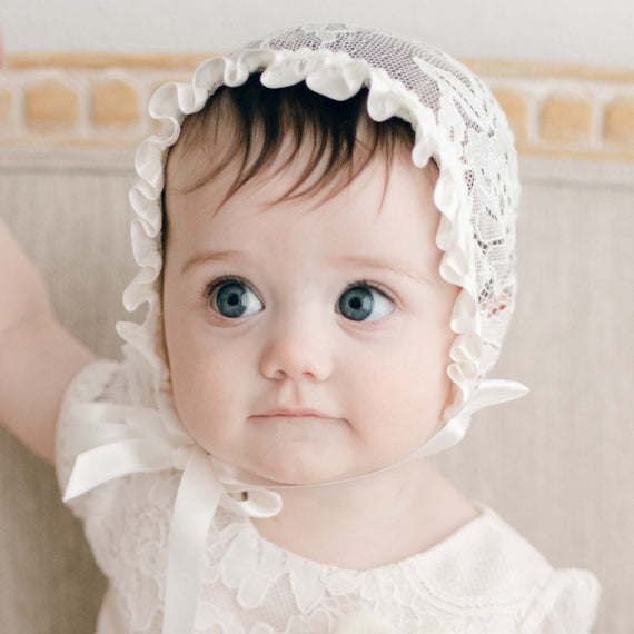 A baby girl with big blue eyes looks to the side with slight smirk. She is wearing the Victoria Lace Bonnet and matching Victoria Lace Dress. The bonnet made with ivory floral lace and ivory silk ribbon is tied snug over her head.