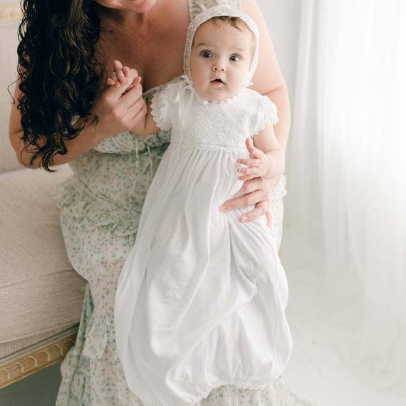 A newborn girl wearing the Ella Layette gown and bonnet is held by her mother.