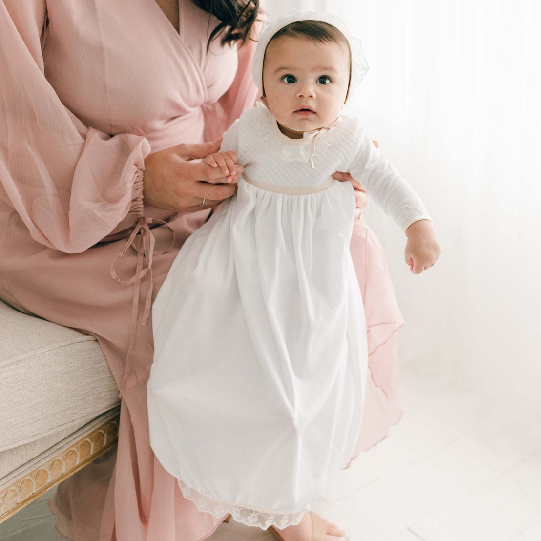 Baby girl wearing the Tessa Quilt Newborn Gown accompanied by her mother. The newborn gown is made with soft pima cotton in white and features a plush white quilt bodice with an ivory Venice lace accent along the neck and cuffs.