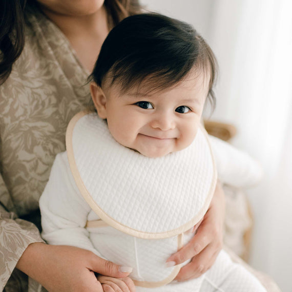 Baby boy smiling and wearing the Liam Boys Bib crafted from soft quilted cotton in ivory and detailed with a silk champagne trim 