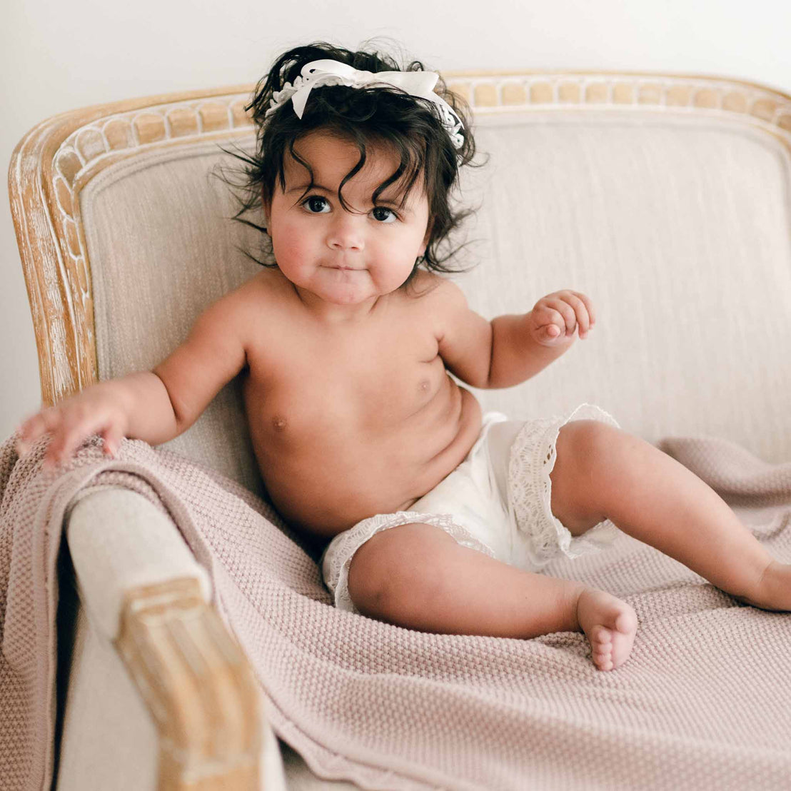 Baby sitting on a chair and wearing the Lily Bloomers made with 100% light ivory cotton, light ivory lace, and ivory silk ribbon