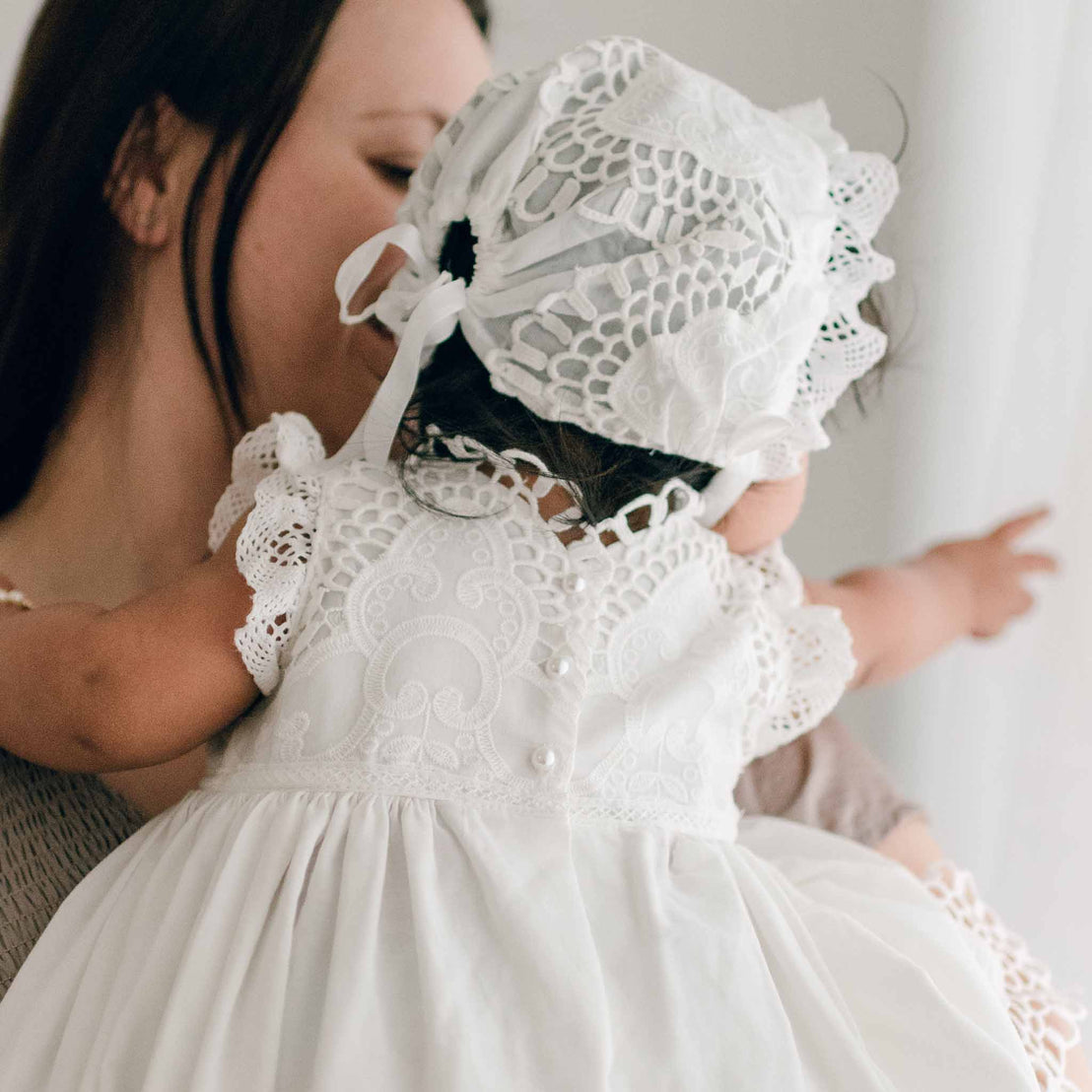 Back view of a baby wearing the Lily Bonnet. The baby sits in her mom's arms and points to something off to the side. The photo shows the details on the back, including the ivory silk ribbon.