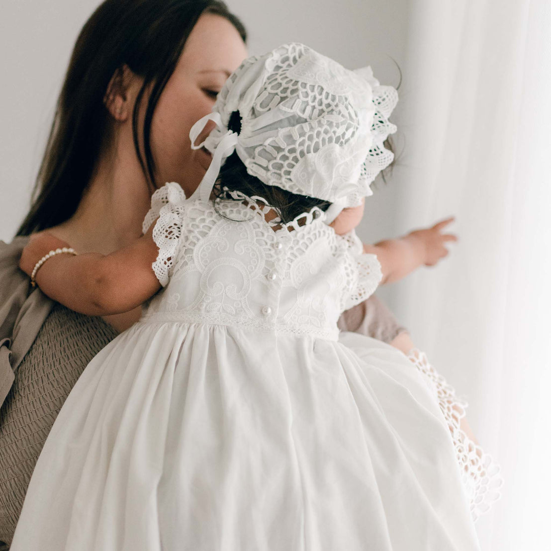 Photo of a baby wearing a Lily Christening Gown & Bonnet. She is held by her mom and the photo showcases the back of the bodice and bonnet designs