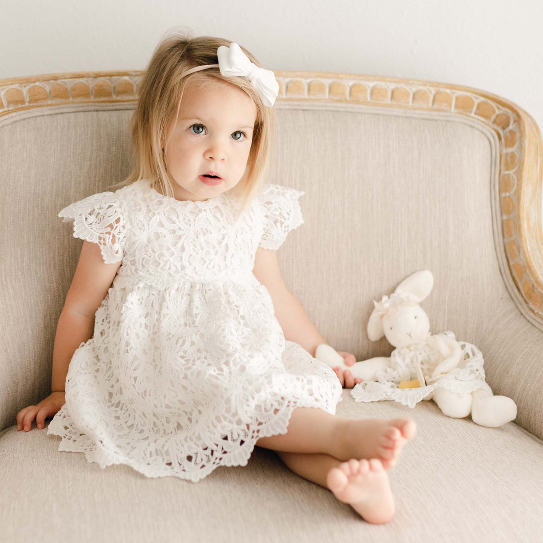 Toddler girl sits on a large upholstered linen chair and wears the Lola Dress and velvet headband. In her left hand is  the Lola Silly Bunny Buddy pacifier holder with a matching dress.